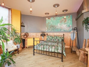 A room with a bed, wooden floor, hanging lights, a fan, a chair, and a large abstract painting on the wall. - Mermaid's Rest, Cornwall 1 bed (Newquay)