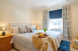 A neatly made bed with pillows, a bedside lamp, a tray with fruits and a vase of flowers, and a window with curtains. - 17 Solent Landing, Bembridge (Bembridge)