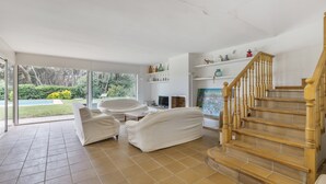 Amplia sala de estar con un conjunto de sofás cubiertos, una escalera de madera y una piscina visible a través de grandes puertas de cristal.