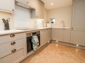 A kitchen with light-colored cabinets, a built-in oven, a sink with a gold faucet, and a window with a plant. - Henside Cottage, Settle (Settle)