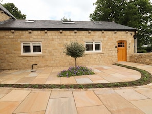 A stone building with a small tree and purple flowers in front, a curved stone pathway, and a wooden door. - Heron View, Longridge (Preston)