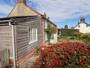 Une maison en bois rustique, dotée d’une cheminée, d’une fenêtre et d’un jardin où poussent des baies rouges.