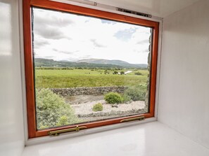 Ein Fenster mit Holzrahmen und Blick auf ein Wiesenfeld mit Steinmauer und kleinen Büschen.