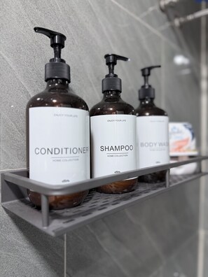 Trois bouteilles d’après-shampoing, de shampoing et de gel douche sont posées sur une étagère en métal dans une salle de bain.