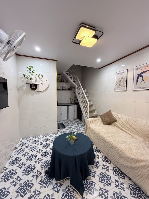 Un salon moderne avec un sol à motifs, une table ronde ornée d’un vase, un canapé, un escalier et un ventilateur fixé au mur.