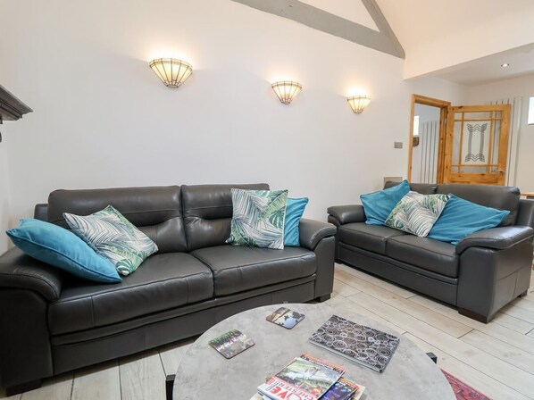 A living room with two black leather sofas, each with patterned cushions, a coffee table with magazines, and wall-mounted light fixtures. - The Stables is in Chester (Chester)