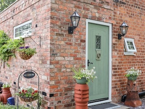 A brick building with a green door, two wall-mounted lanterns, and hanging flower baskets. - The Stables is in Chester (Chester)