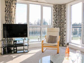 Ein Wohnzimmer mit Fernseher, einem Glastisch, einem Sessel und Blick durch große Fenster.