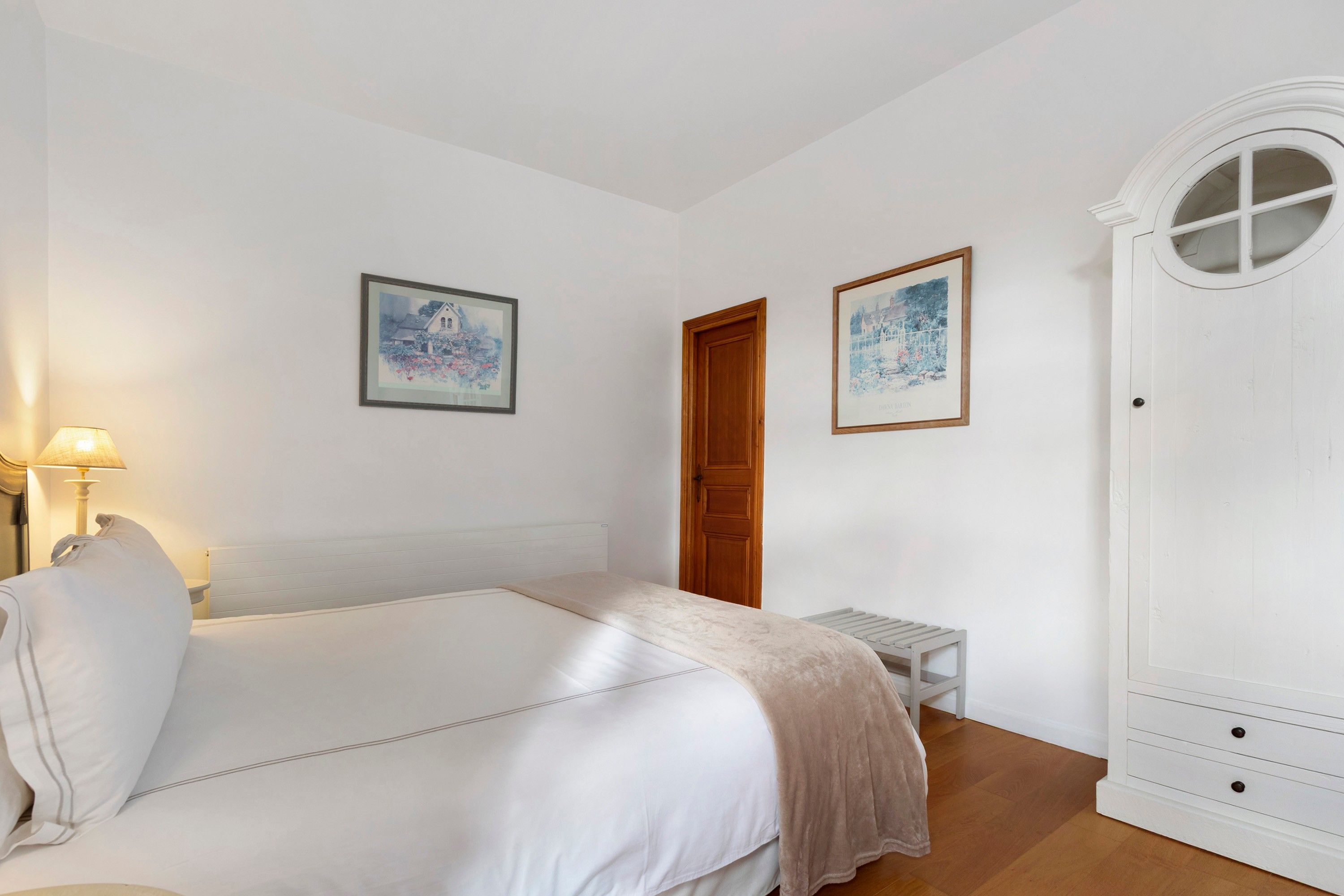 A bedroom with a bed, a bedside table, a lamp, a wardrobe, and two framed pictures on the wall.