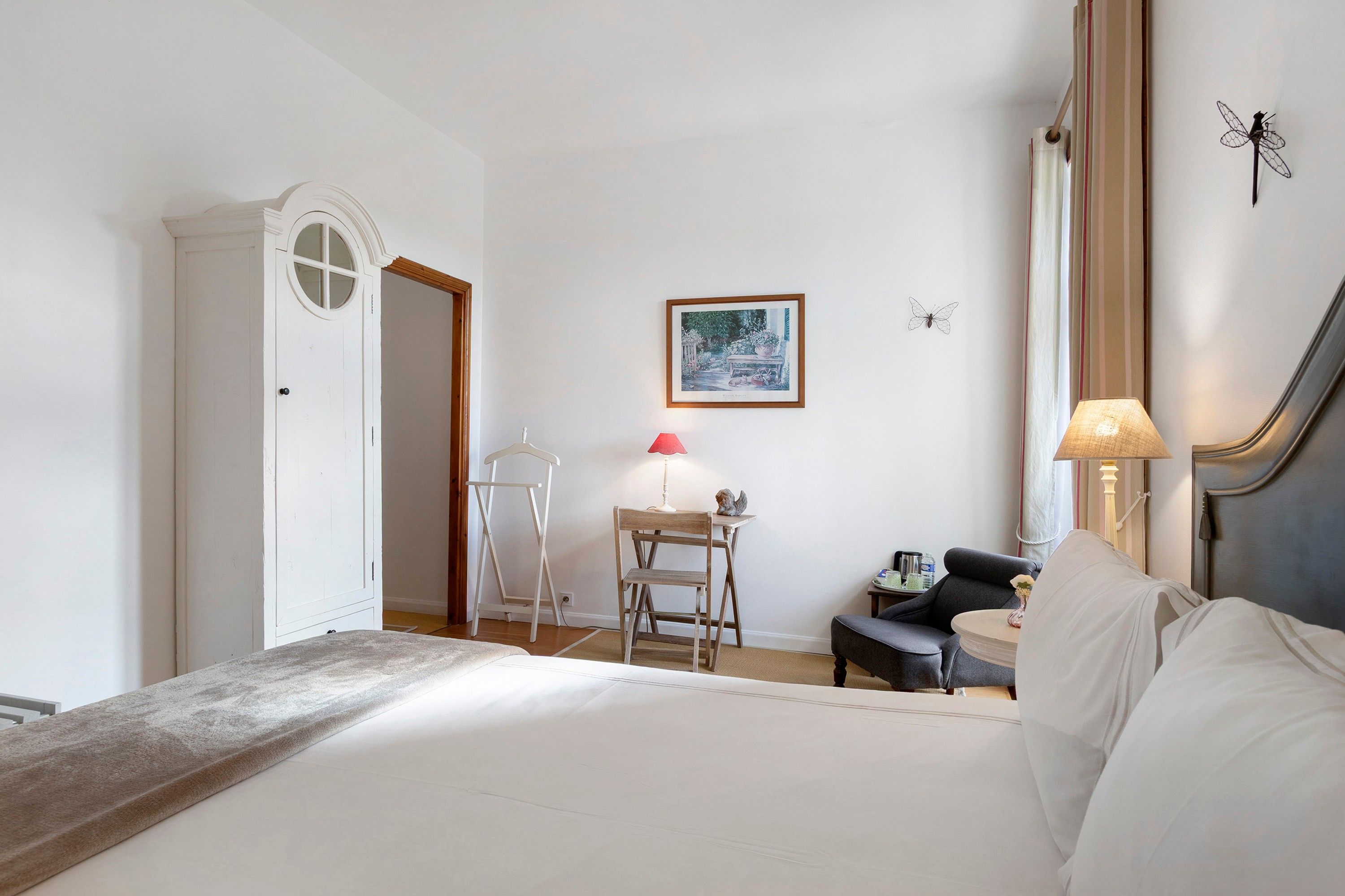 A hotel room with a bed, a desk, a chair, a lamp, and a framed painting on the wall.