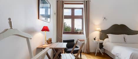 A bedroom with a bed, a desk with a chair, a window with curtains, and a nightstand with a lamp.