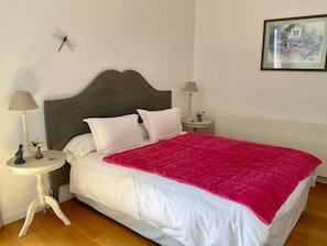 Un letto rifatto con cura, una coperta rossa, due comodini con lampade, un'opera d'arte appesa al muro e un piccolo oggetto decorativo sul tavolo a sinistra.