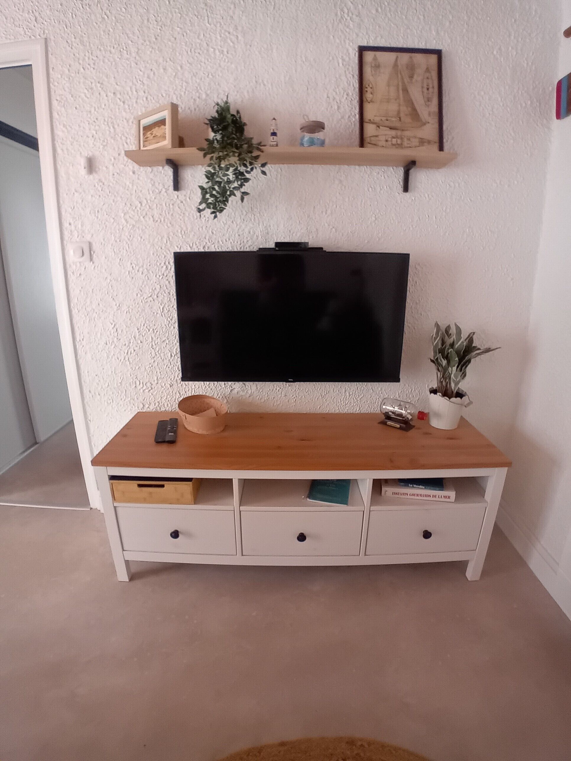 Ett rum med en tv monterad på en vägg med en intressant yta, ett träbord med lådor, en hylla ovanför tv:n med dekorativa föremål och en krukväxt till höger.