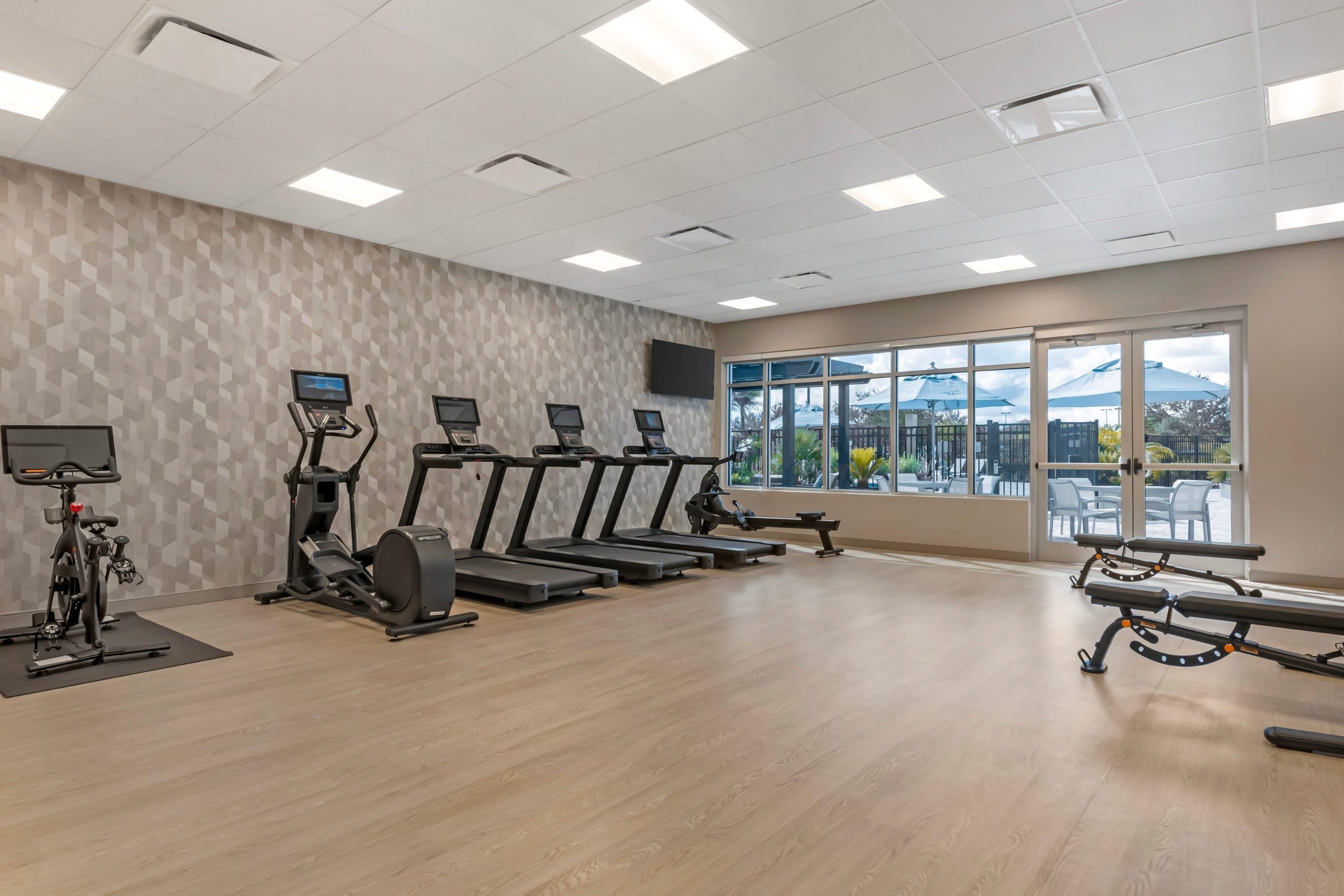 A gym with treadmills, an elliptical, and a stationary bike, with a large window offering a view of the outdoors.