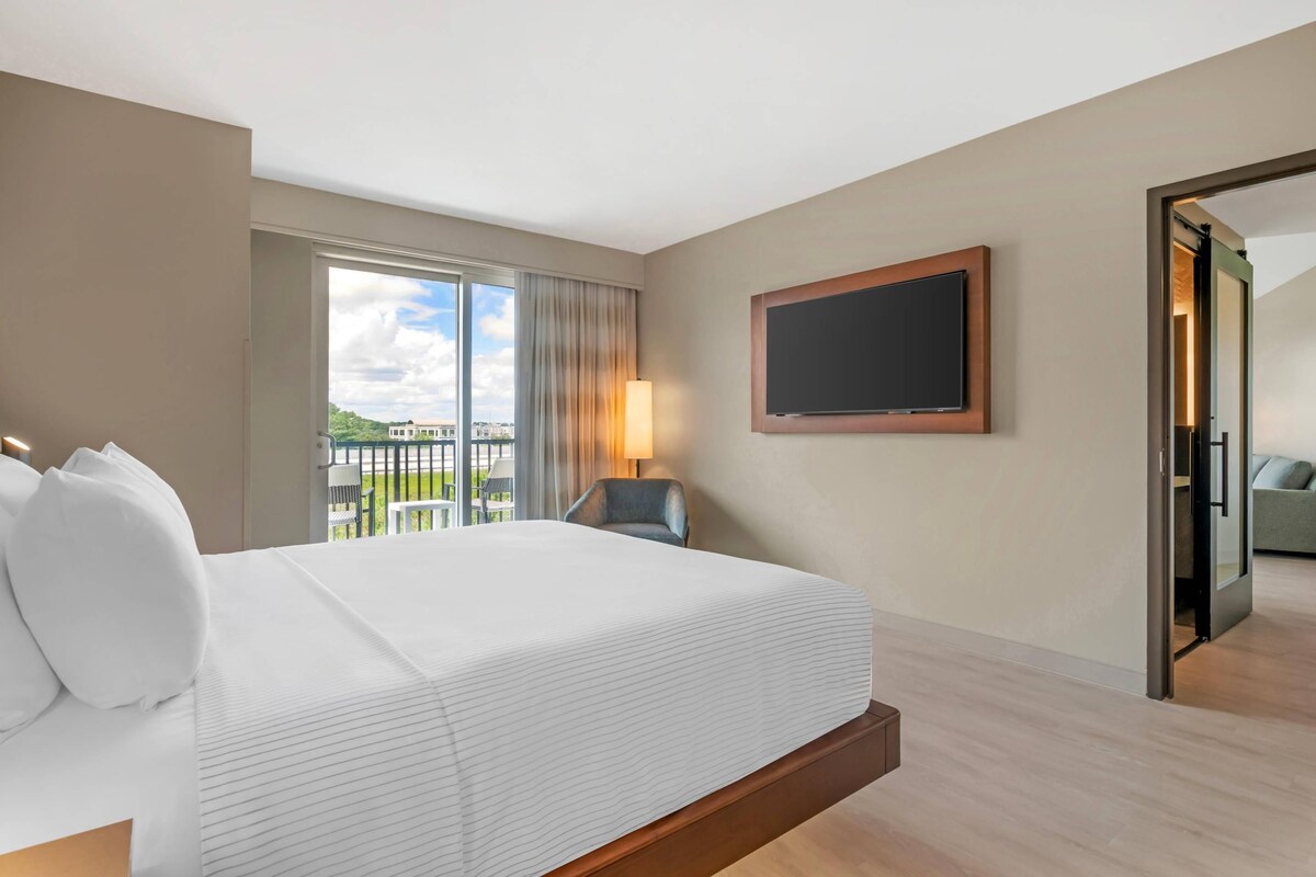 Quarto de hotel com cama grande, televisão de tela plana na parede, varanda com vista e uma cadeira.