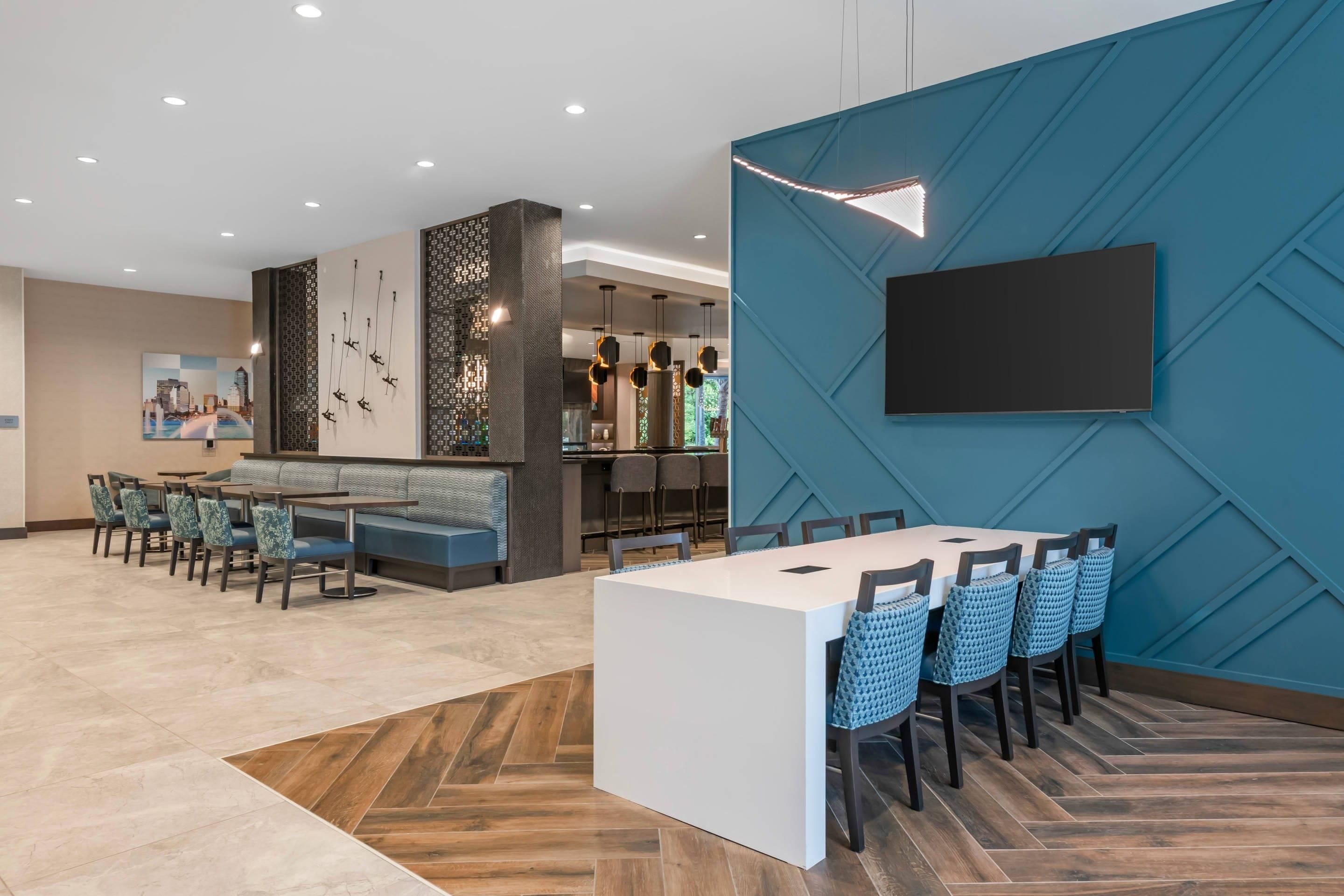 A modern dining area with a long table and chairs, a bar area with seating, and a large TV mounted on a blue wall.