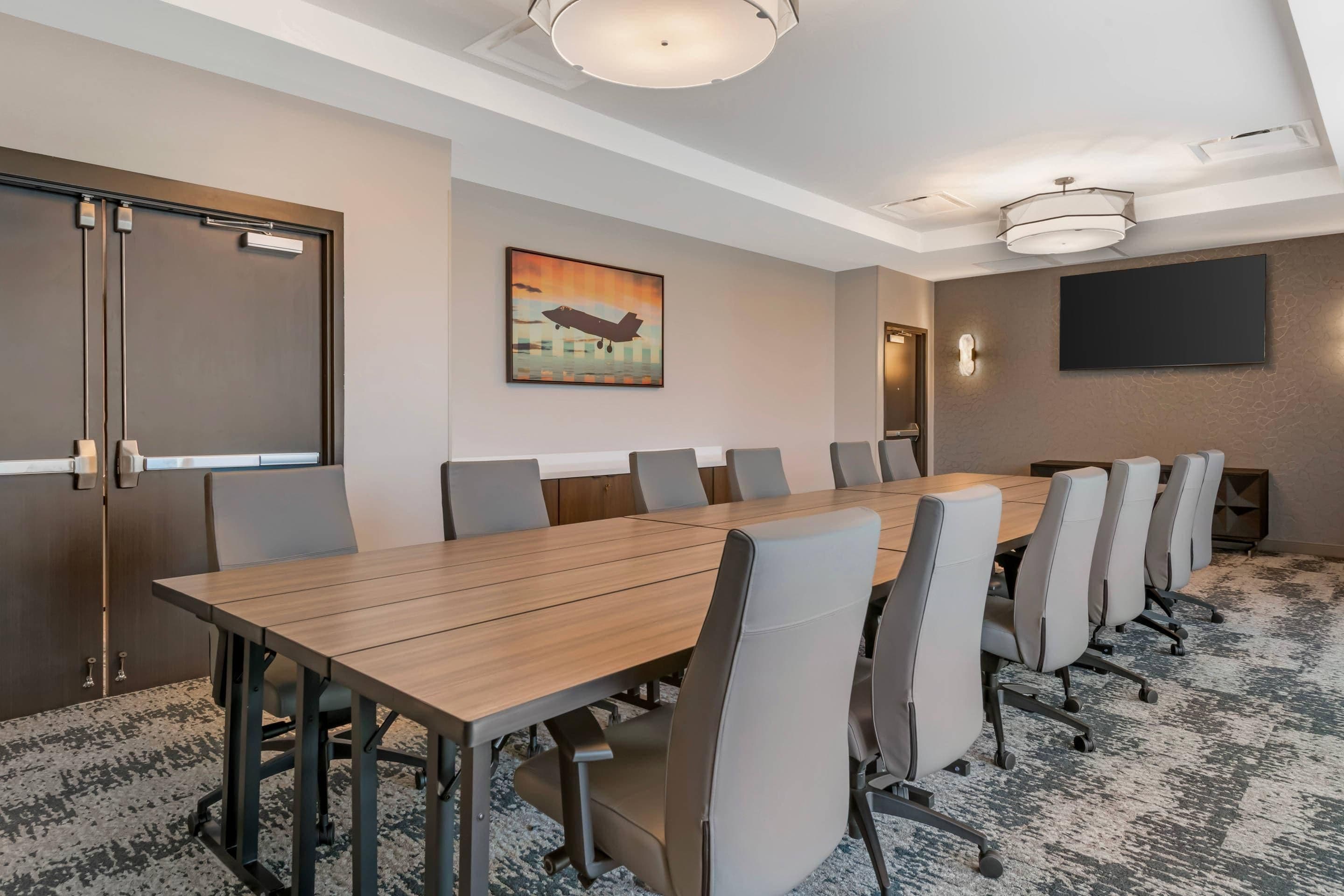 A conference room with a long table, multiple chairs, a painting on the wall, a flat-screen TV, and a door with a handle.