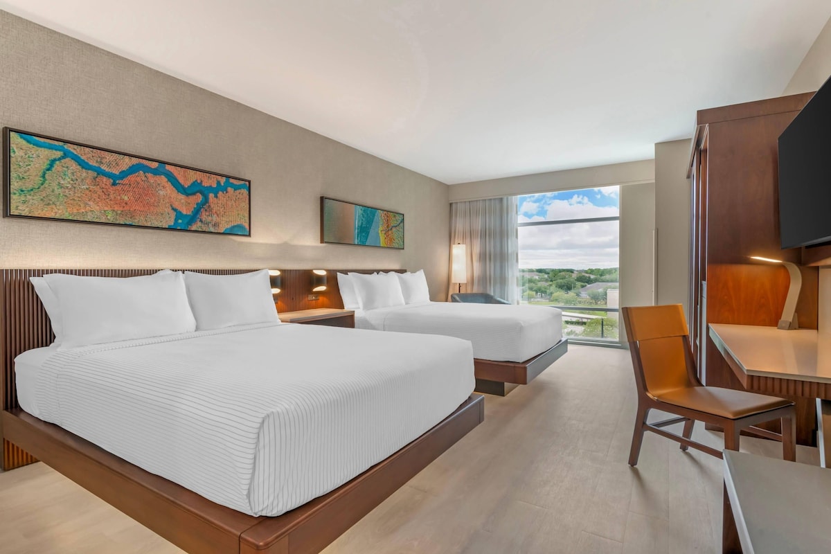 Ein Hotelzimmer mit zwei Betten, einem Schreibtisch, einem Sessel, einem Fernseher und einem großen Fenster mit Blick.