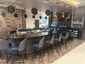 A modern bar area with a curved bar, bar stools, and pendant lighting. - Cambria Hotel Jacksonville (Jacksonville)