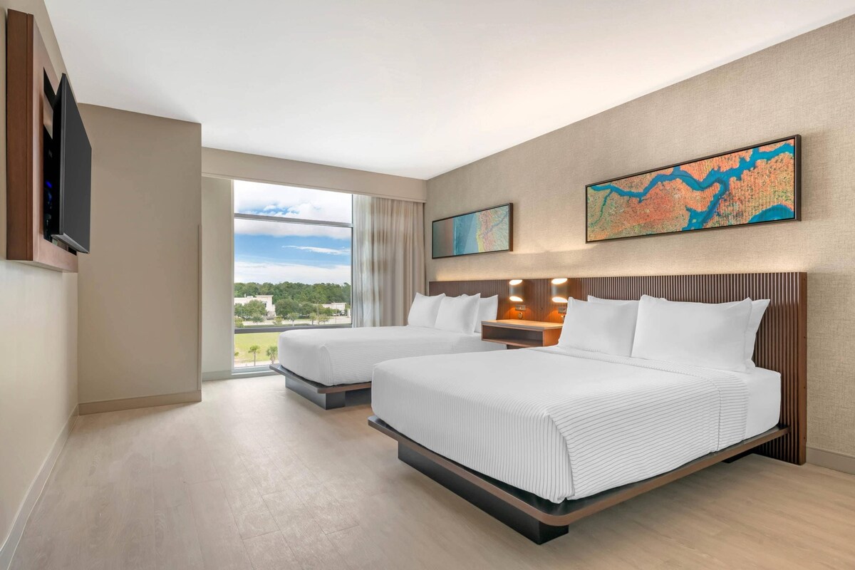 Quarto de hotel com duas camas, uma janela grande com vista e duas obras de arte penduradas na parede.