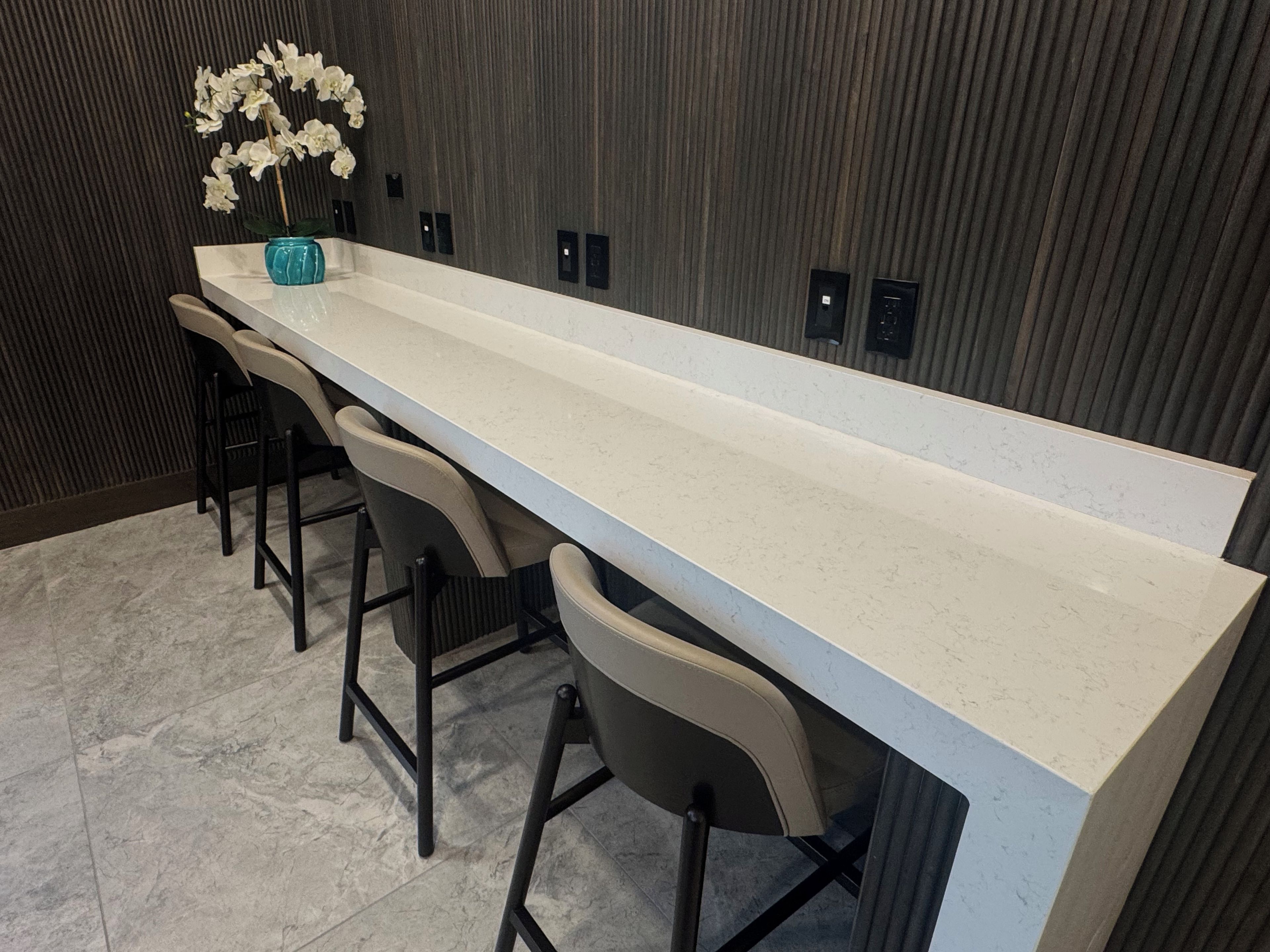 Una cocina alargada y estrecha con encimera blanca, cuatro taburetes de bar y un florero con flores blancas a la izquierda.