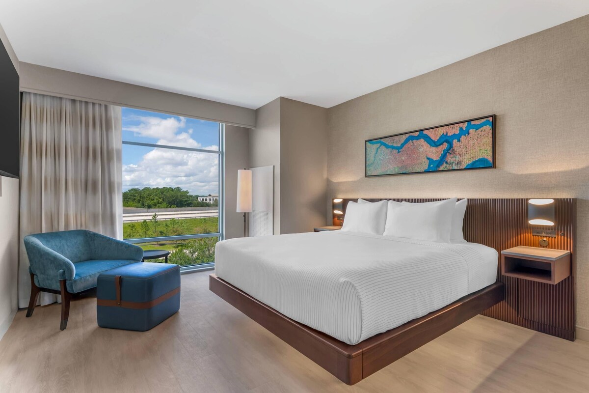 Quarto de hotel com uma cama grande, área de estar com uma poltrona, escrivaninha e uma janela ampla com vista para áreas verdes e uma estrada.