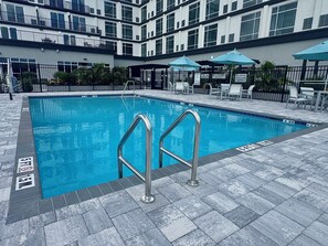 A swimming pool with metal ladders, surrounded by a tiled deck and outdoor furniture. - Cambria Hotel Jacksonville (Jacksonville)