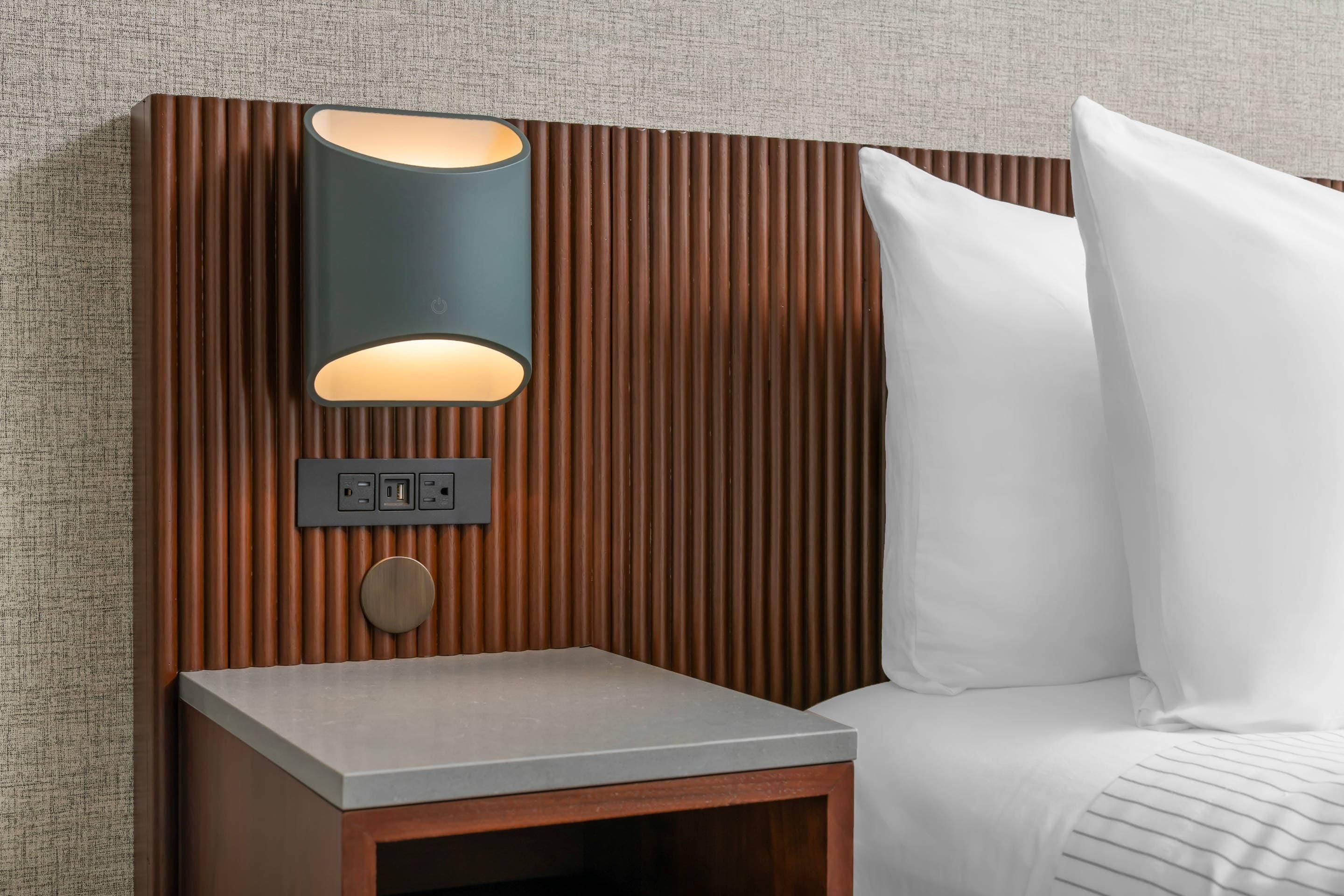 A modern bedroom with a wooden headboard, a bedside table, a lamp, and a white pillow.