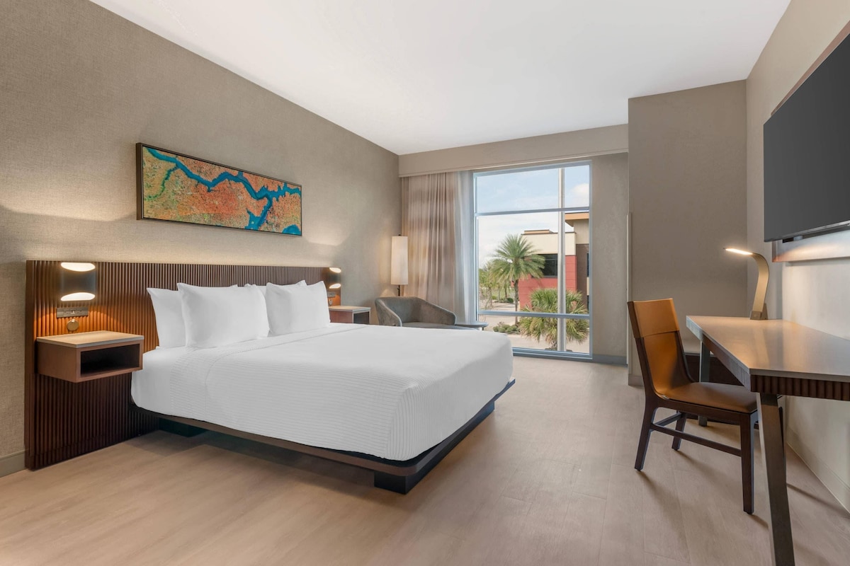 Quarto de hotel com uma cama grande, uma escrivaninha, uma cadeira, uma televisão e uma janela com vista para palmeiras.