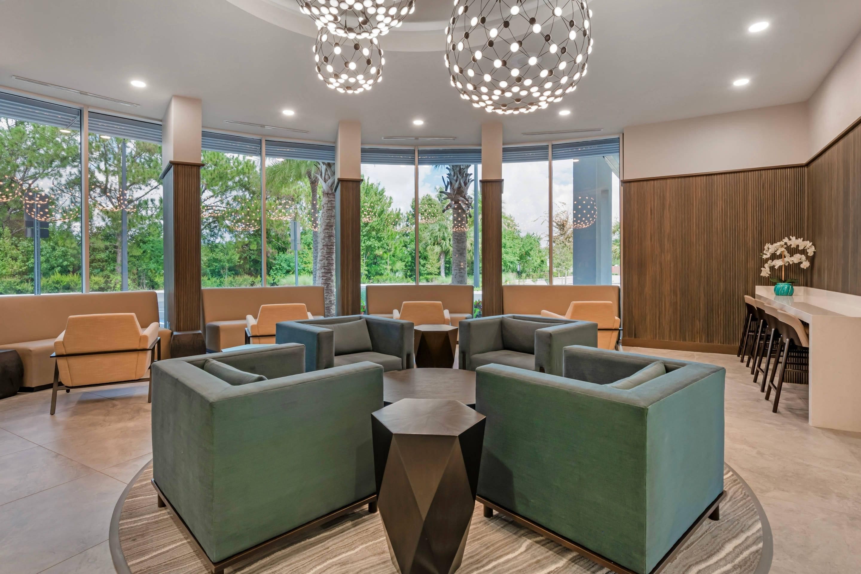 A modern lounge area with green sofas, a central coffee table, and a bar with stools. Large windows provide a view of the outdoors.