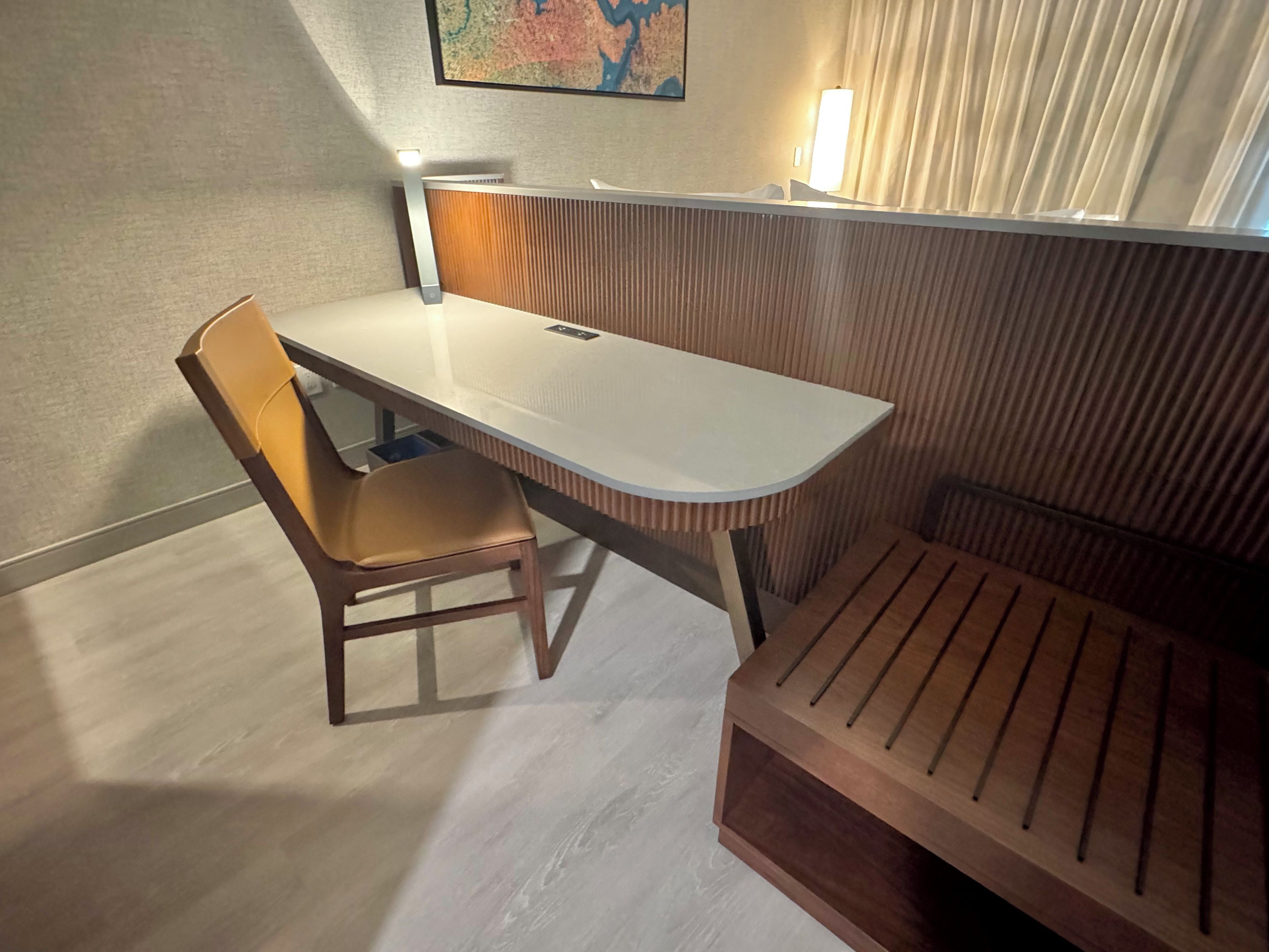 A room with a desk, a chair, a bench, a map on the wall, and a window with curtains.