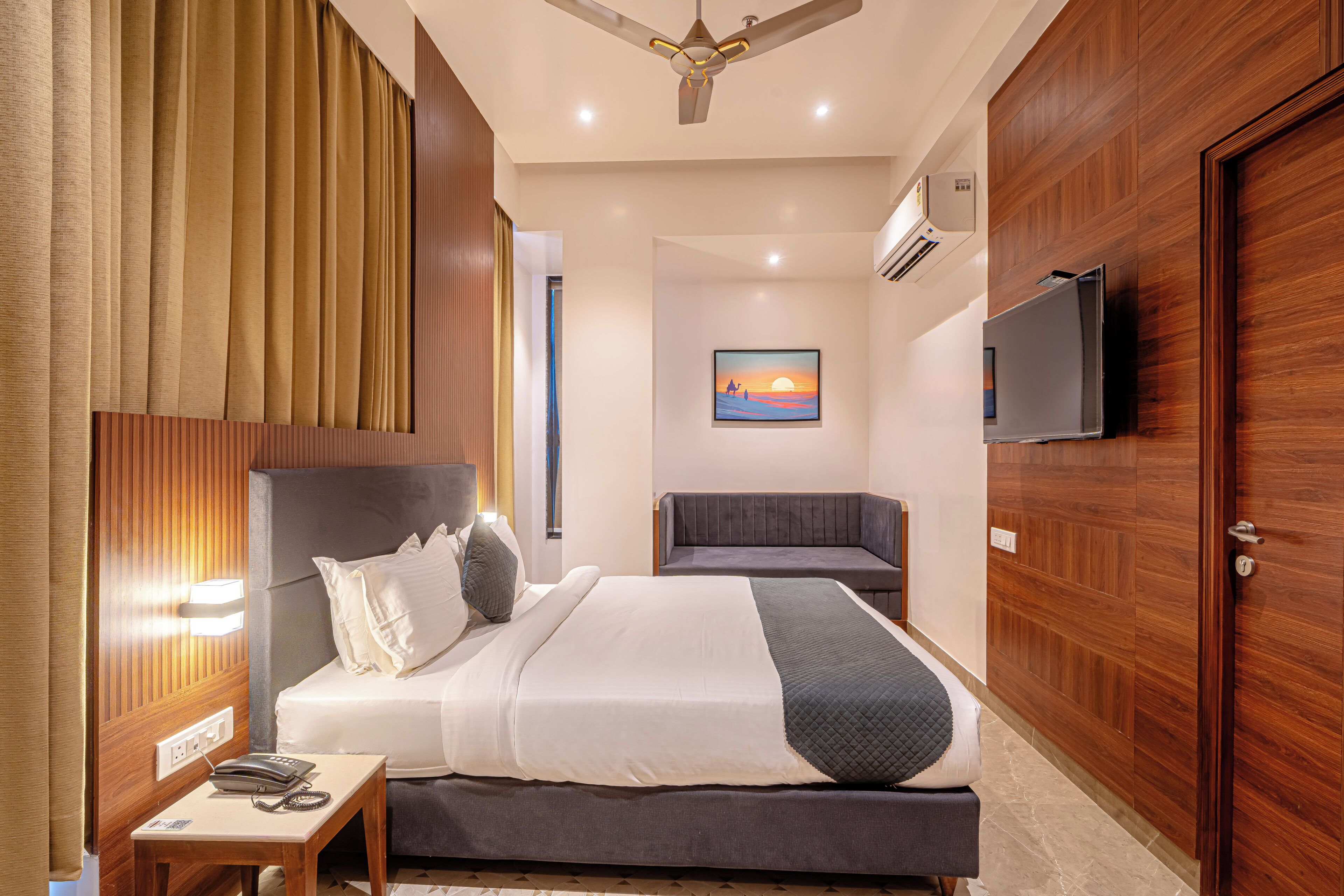 A hotel room with a bed, bedside table, lamp, TV, and a painting on the wall.