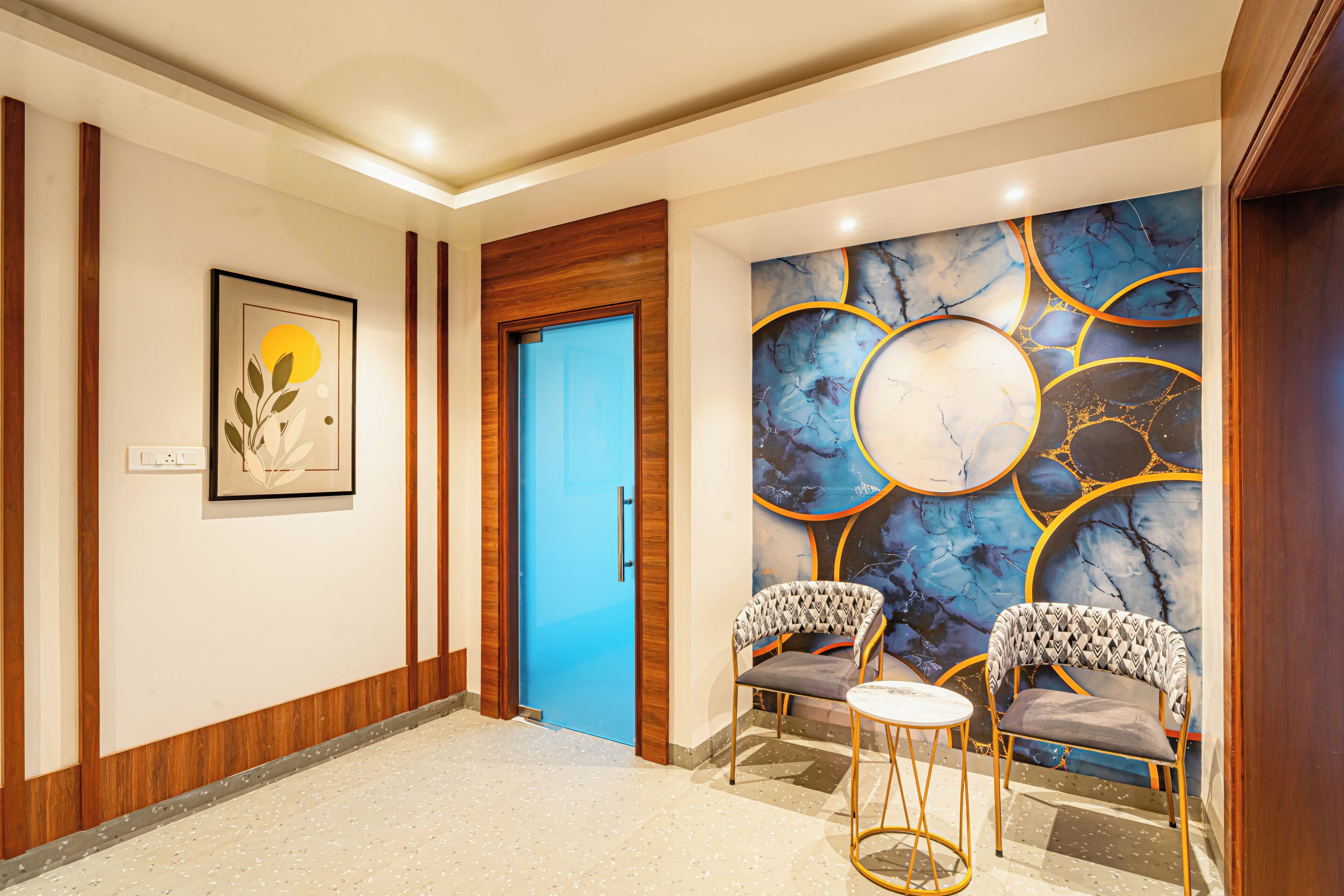 Un vestíbulo de hotel con una puerta azul, una obra de arte enmarcada, una pared de mármol con diseños circulares y dos sillones estampados.