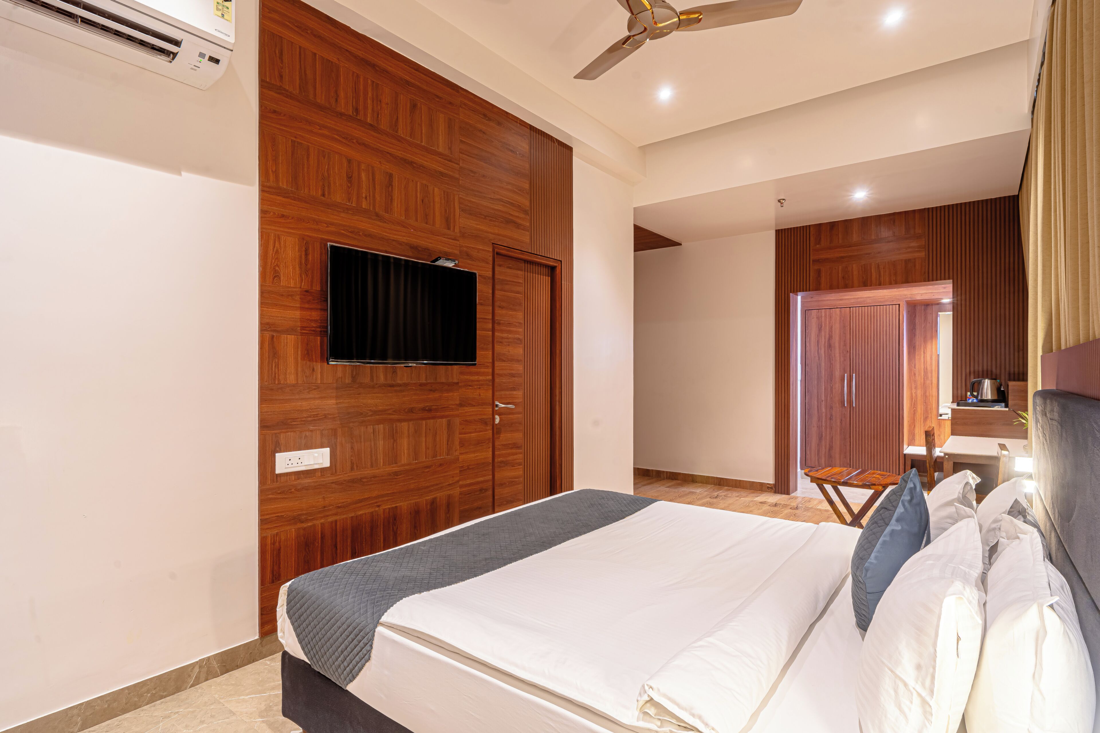 A bedroom with a bed, a TV mounted on a wooden wall, a ceiling fan, and a wooden wardrobe.