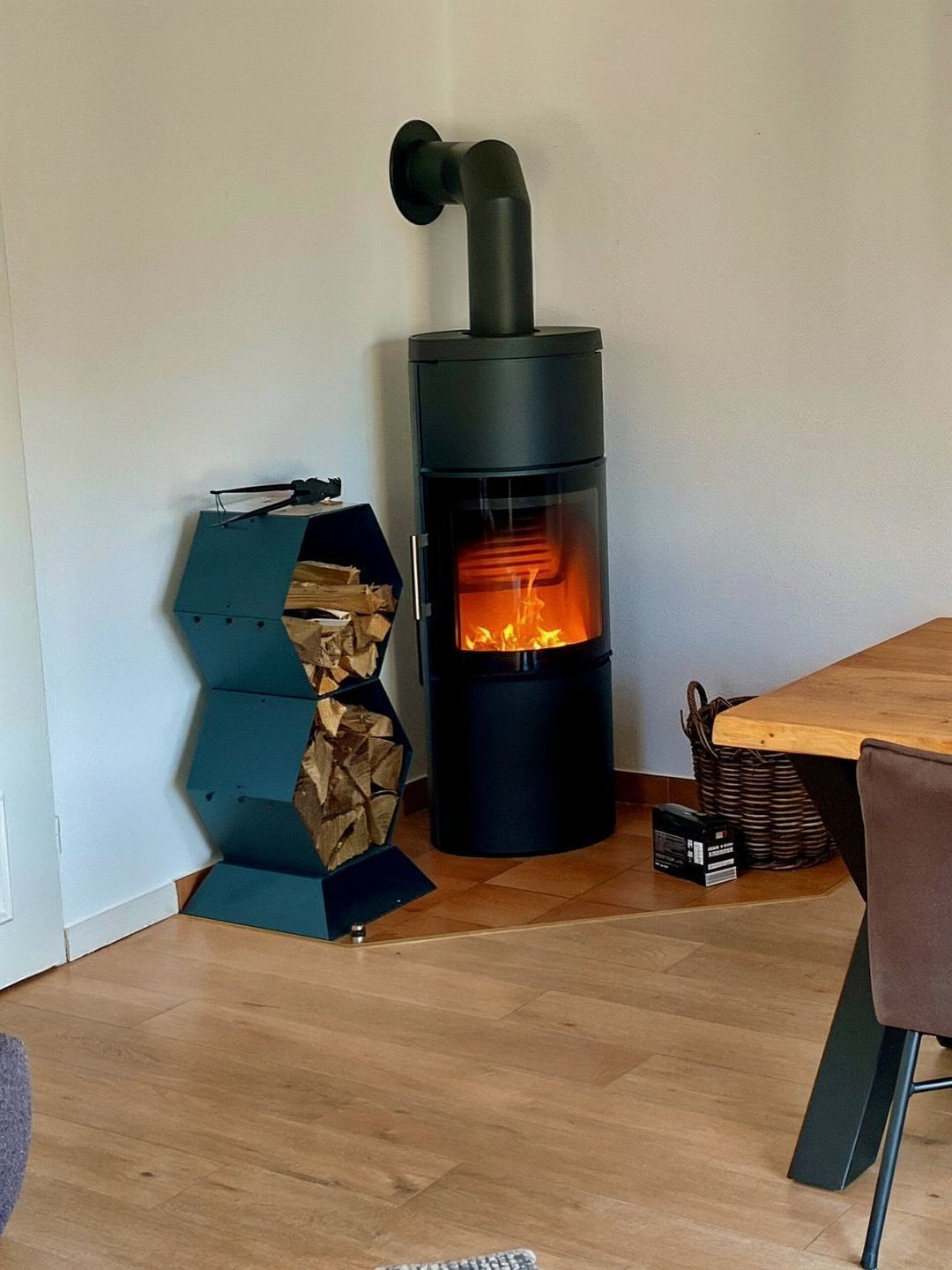 Een moderne houtkachel met glazen ruit, een blauwe opbergunit, en een eettafel met een stoel.