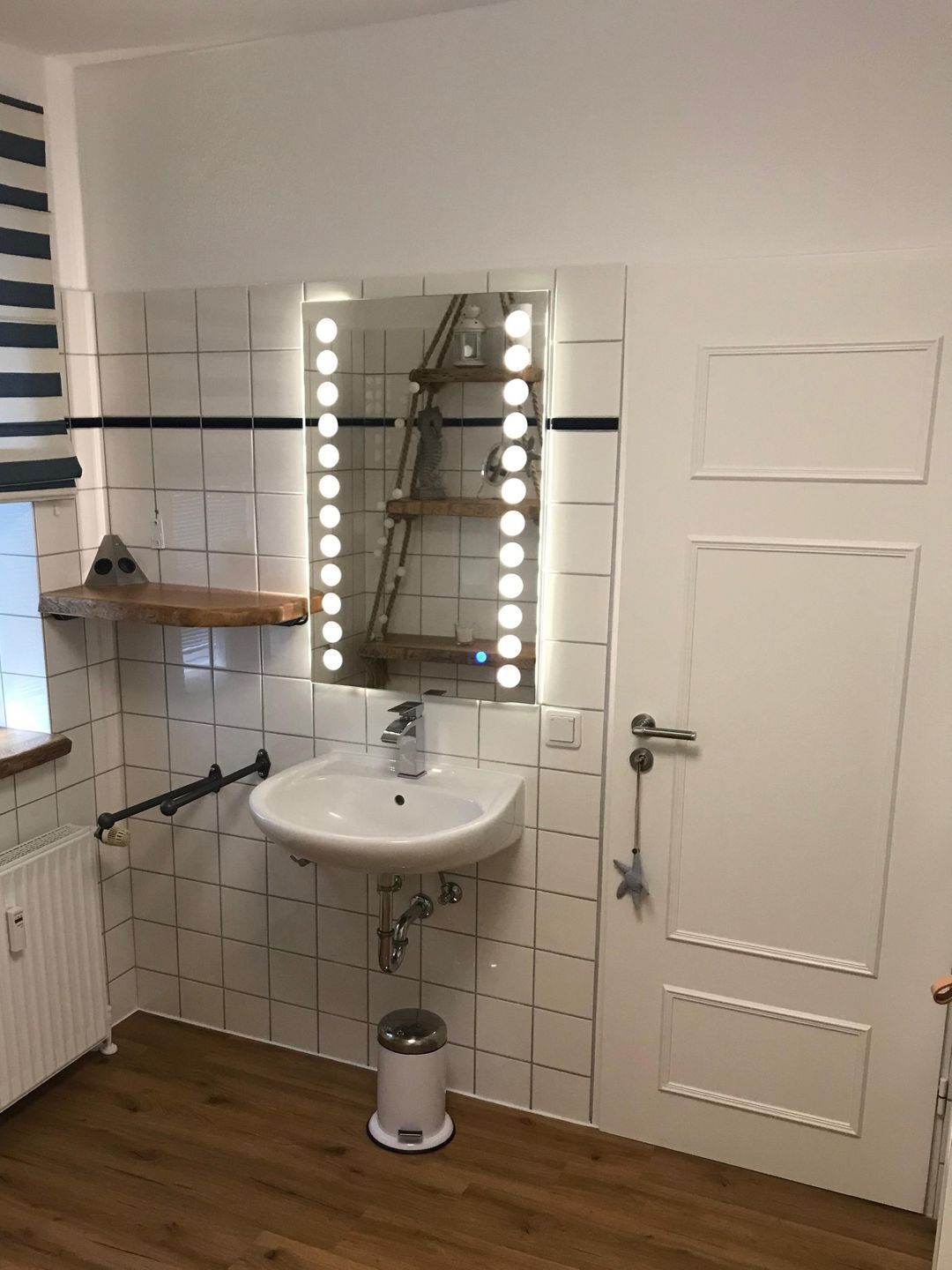 A bathroom with a white sink, mirror with lights, wooden floor, and white tiled walls.