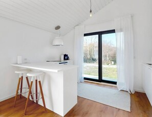 A modern kitchen with a white countertop, bar stools, a hanging light, and a large window with a view of trees. - Stylish Cottage Near Beach, Cliff and Harbour Life (Rodvig Stevns)