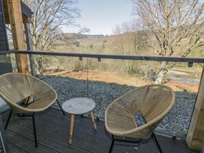 Eine Holzterrasse mit zwei Korbstühlen und einem kleinen runden Tisch, mit Blick auf eine ländliche Landschaft mit Bäumen und Hügeln.