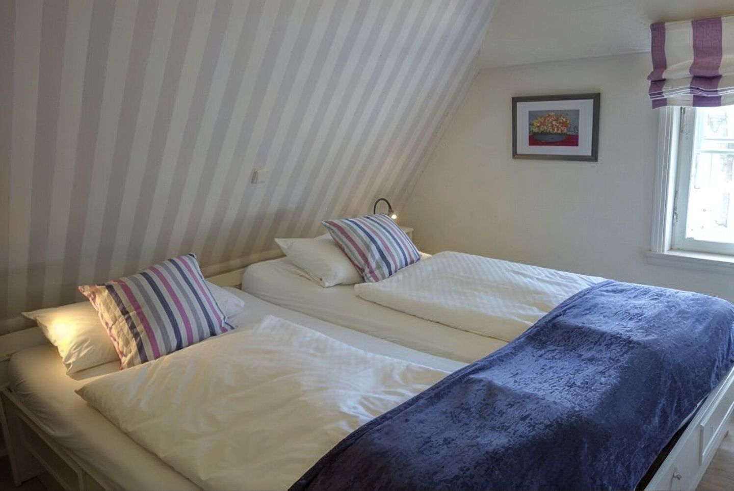 Une chambre avec deux lits, des rideaux à rayures, un tableau encadré au mur et une fenêtre également dotée de rideaux.