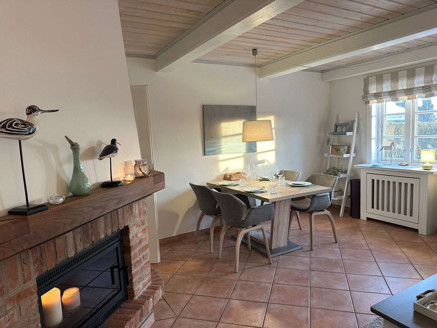 Zona de dinar amb una llar de pedra, taula preparada per a quatre i finestra amb cortines.
