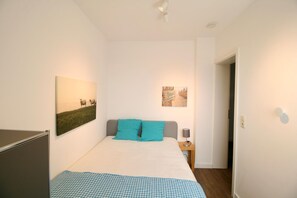 Un dormitorio con una cama, dos almohadas, una mesita de noche, una lámpara y dos cuadros en la pared.