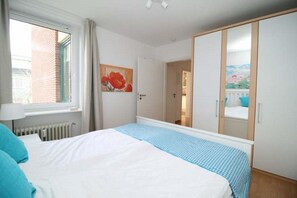 A bedroom with a bed, a window, a radiator, a wardrobe, and a painting. - Strandmohn - Holiday apartment Strandmohn: 60 m² on the beach with balcony & sea view (Wyk auf Föhr)