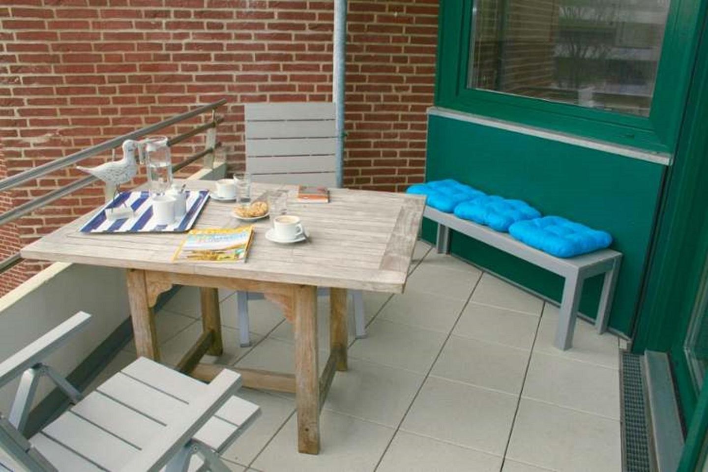 Ein Holztisch mit einer Zeitschrift, einer Tasse und einem Glas Wasser auf einem Balkon mit einer Ziegelwand und einem grünen Fensterrahmen.
