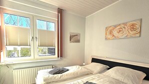 A bedroom with a bed, a radiator, a window with blinds, and a painting on the wall. - Austernfischer - Holiday apartment Austernfischer in Wyk auf Föhr - close to the beach with (Wyk auf Föhr)