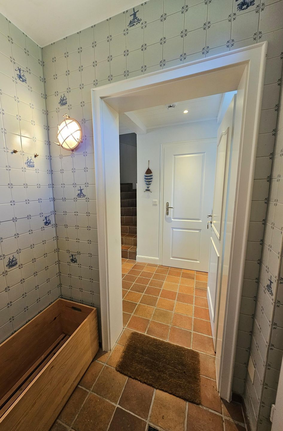 A hallway with patterned wallpaper, a wooden bench, a lit wall sconce, and a door leading to another room.