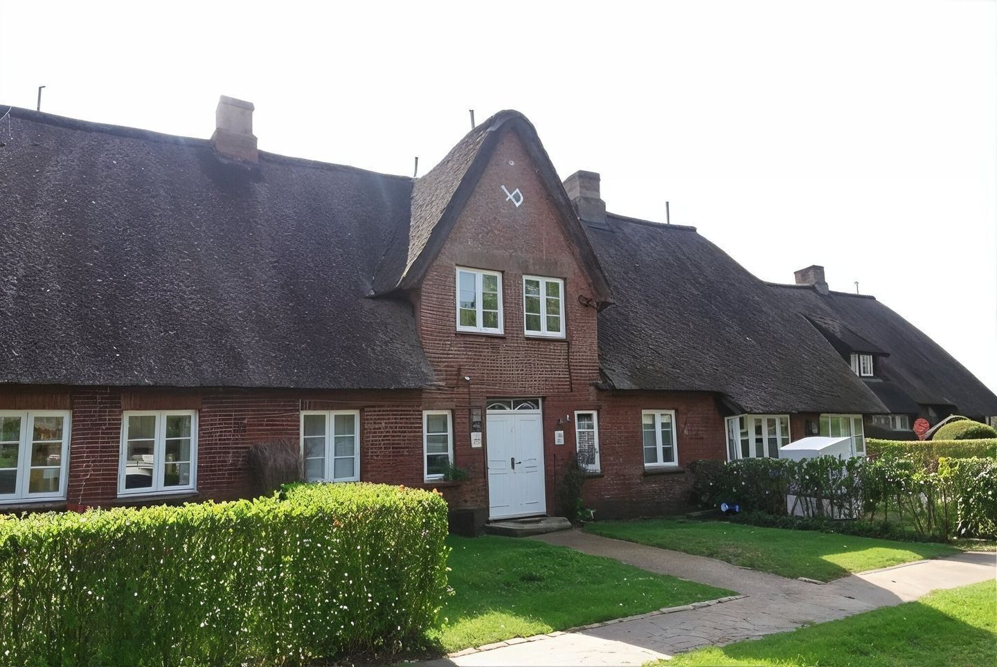 A traditional brick building with a steeply pitched thatched roof, a central entrance with a white door, and a small garden area with a hedge and a pathway leading to the entrance.