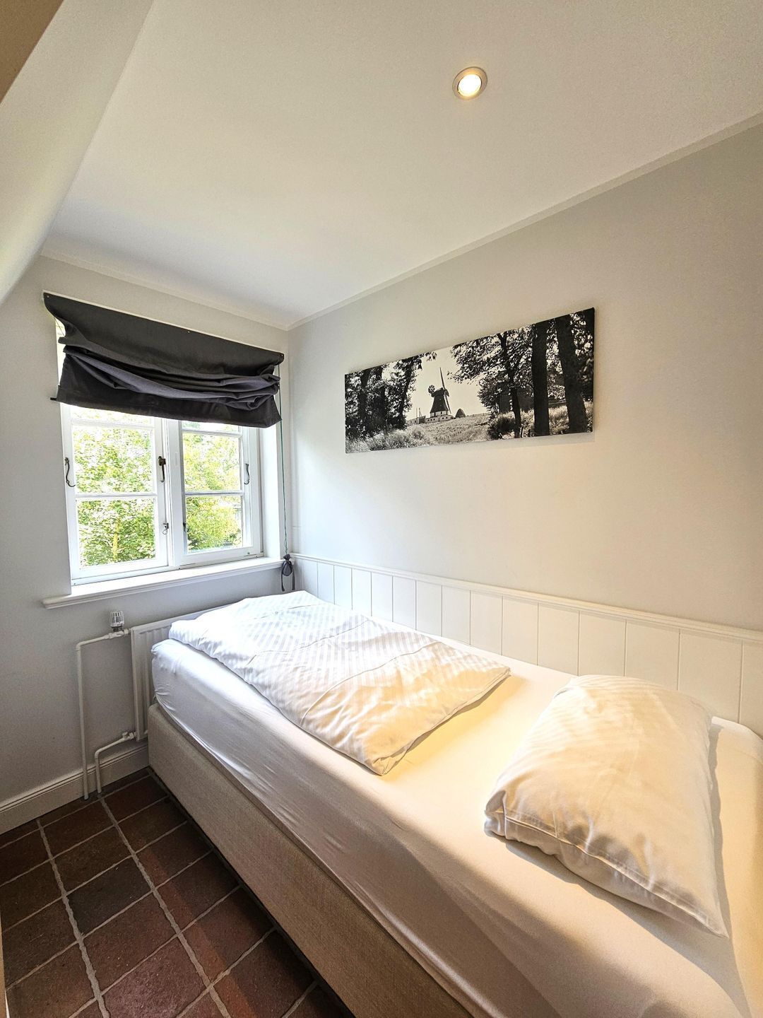 A single bed with white bedding, a window with a black valance, a radiator, and a black and white artwork on the wall.