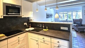 A modern kitchen with white cabinets, a built-in oven, a black stovetop, pendant lights, and a view of a living area with a chair. - Pastoratshof - House 6 - 5-star vacation home Pastoratshof - House 6 with sauna & sauna facilities (Wrixum auf Föhr)