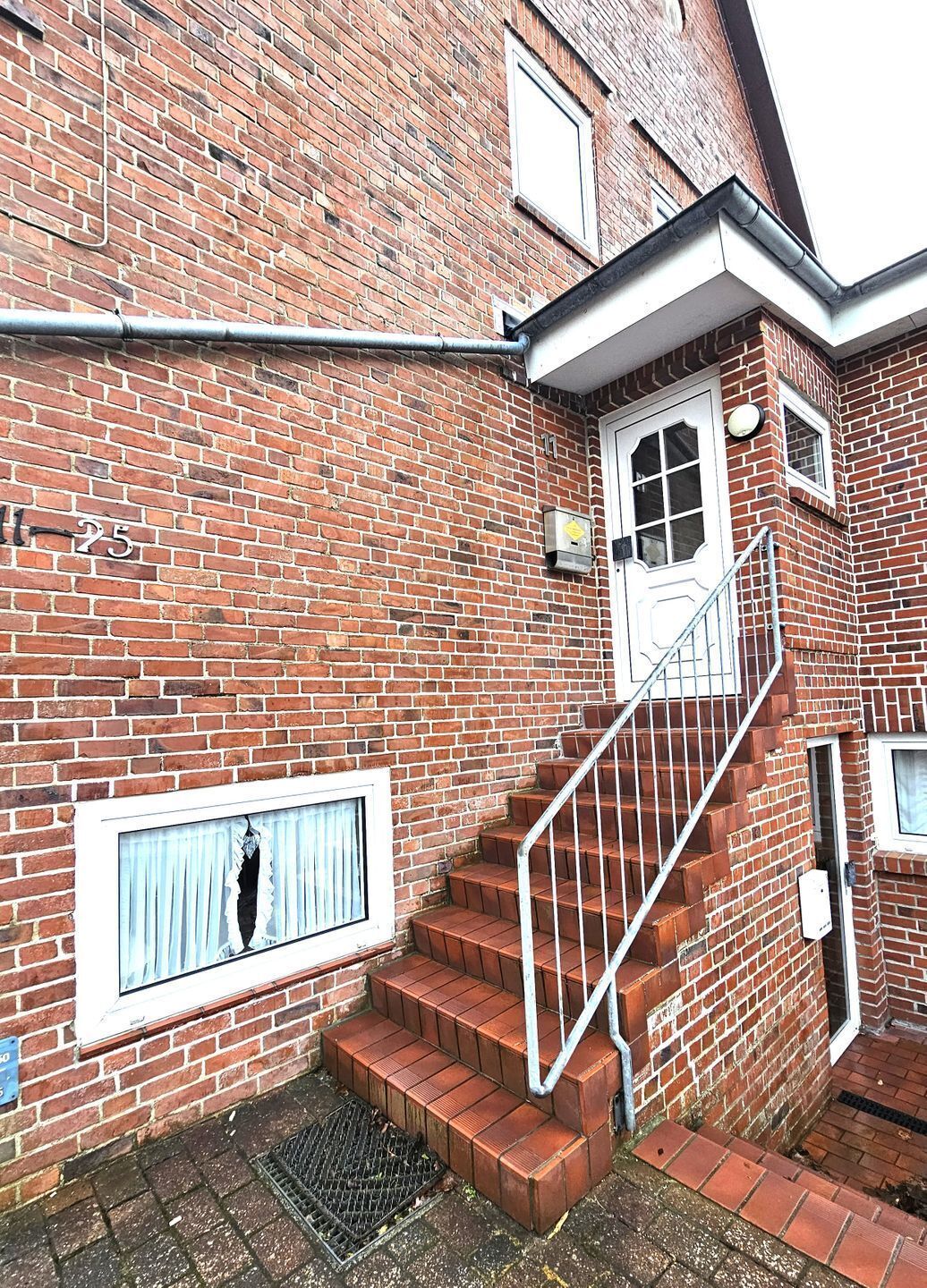 Ein Backsteinhaus mit einer weißen Tür, einem Fenster mit Vorhang und einer Metalltreppe.