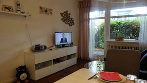 Ein Wohnzimmer mit Fernseher, einem Esstisch für zwei Personen, einer Couch, einem Wandbild und einem Fenster mit Blick auf Grünflächen.